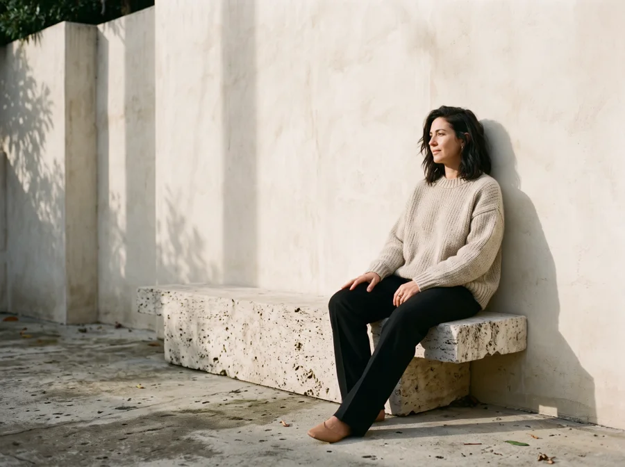 A woman in an oatmeal knit and dark trousers in a sunlit architectural setting